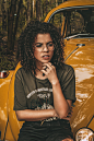 A young woman in glasses seated on the hood of a yellow Volkswagen Beetle