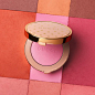 A still life shows an open Gucci Blush de Beauté – Powder Blush, displaying a pastel pink powder. The background is a chequered print of the different powder shades available.