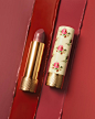 A still life features an open Gucci Rouge à Lèvres Voile – Sheer Lipstick lying next its floral yellow case. The background is four different streaks of red and pink lipstick to demonstrate the different shades in the collection.