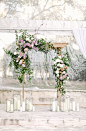 Elegant wedding arch. Gorgeously decorated natural wood trellis covered in overgrown wild and romantic greenery, vines, and pink, purple, and white flowers. Made romantic by adding tall pillar candles in varying sizes of glass vases. Super dreamy for a sp