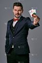 身着黑色经典西装和红色领结的男子展示了两张扑克牌和筹码，在灰色的工作室背景下摆姿势。赌博、扑克、赌场