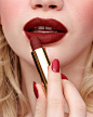 A close up shows a blonde woman applying a deep red Gucci Rouge à Lèvres Mat – Matte Lipstick to her lips, displaying matching red nails. Her skin is translucent against the dark red shade of lipstick.