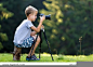 Young blond child boy sitting on tree stump on grassy clearing taking picture with tripod camera.