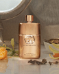 A bottle of the gold Gucci Guilty Eau de Parfum Intense Pour Femme stands on a table. The Gucci logo catches the light and sparkles, sprinkled around it lie part of an orange, spices in a bowl and other ingredients from the fragrance.