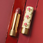 A still life features an open Gucci Rouge à Lèvres Voile – Sheer Lipstick lying next its floral yellow case. The background is four different streaks of red and pink lipstick to demonstrate the different shades in the collection.