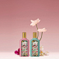 Against a pink blush background, a still life shows a bottle of the pink Gucci Flora Gorgeous Gardenia next to the green bottle of the Gucci Flora Gorgeous Jasmine. A fuchsia flower appears on behind Gucci Flora Gorgeous Gardenia, while a tall blooming wh