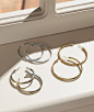 A color photograph shows three pairs of David Yurman women’s hoop earrings from the Cable Collection scattered on a white windowsill with hard shadows. The jewelry is crafted from Cabled 18K yellow gold or sterling silver with or without 18K yellow gold a