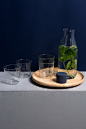 Rivington玻璃器皿系列的正视图，四个不倒翁，其中两个被堆叠在一起，一个玻璃水瓶带有蓝色硅胶塞，位于其旁边。 玻璃水瓶内是水和薄荷叶。 玻璃水瓶和堆叠的玻璃杯在木板上。 白色的表面和深蓝色背景。