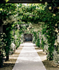 A garden path through an arch leading to an alcove with table and chairs, with climbing plants... by Mint Images on 500px