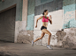 A woman running along an urban street past buildings with peeling paint and a metal shutter.