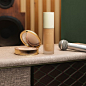 A still life shows an open Poudre De Beauté Mat Naturel Face Powder next to a bottle of Éternité de Beauté foundation and a silver microphone on a grey marble effect table. The background shows a gold and white striped wallpaper, next to a green wall.