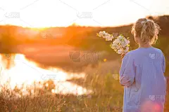 日落时湖边年轻女子的背影。女孩带着苹果树和鲜花在公园里欣赏夏天的日落。快乐的女人捧着花束，在户外。生