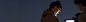 Two people standing outside at night looking at a laptop. There is a blurred city skyline in the background.