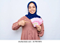 Excited young Asian muslim woman in pink shirt point money banknotes isolated over white background. People religious lifestyle concept 库存照片