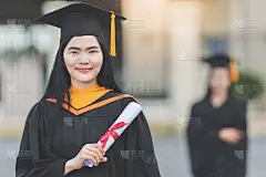 一位年轻的亚洲女子大学毕业生穿着毕业礼服和学士帽，在参加大学毕业典礼后，手持学位证书站在大学大楼前