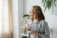 一位年轻女子，留着短发、刘海、眼镜和纹身，手里拿着一杯早茶和茶托，穿着灰色休闲装，站在菜架旁边，种植