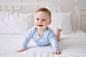 A cute healthy little baby is lying on a bed on white bedding at home in a blue bodysuit the kid looks at the camera smiles