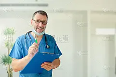 一位慈祥的男医生看着镜头，指着什么东西。他左手拿着一个记事本，右手拿着一支笔。