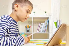 教育、学习、造纸工艺、娱乐。小男孩在家用剪刀剪彩纸，在平板电脑上看视频教程