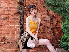 一个快乐的中国女孩的肖像，穿着黄色吊带上衣和短裙看着和微笑的镜头与砖墙背景，无忧无虑的年轻女士享受她