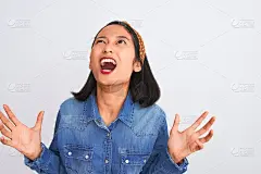 年轻美丽的中国女人穿着牛仔衬衫站在孤立的白色背景疯狂和疯狂的大喊大叫，好斗的表情和手臂举起。挫折的概