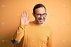 中年男子穿着休闲毛衣，戴着眼镜，孤立的黄色背景挥手问好，快乐和微笑，友好的欢迎手势