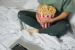 一个小女孩手里拿着爆米花桶，准备躺在床上看电影。好戏上演。吃美味的不健康的甜食。在家看新电影。卧室休