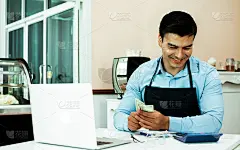 穿着蓝色围裙、戴着眼镜、面带幸福和成功的微笑、手里拿着钱、坐在店里计算利润的白人英俊的咖啡馆老板或企