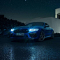 BMW M8 Competition Coupé in Marina Bay Blue metallic with shining LED headlights, standing three-quarter front view in front of villa at night.