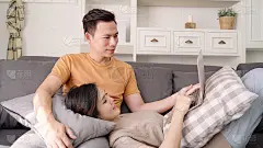 亚洲情侣在家里的客厅里用笔记本电脑搜索网络，甜蜜的情侣在家里放松的时候躺在沙发上享受爱情的时刻。生活