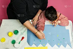 特殊教育的老师和学生穿着长袍，在桌子上用手画着生日花环和涂着丙烯颜料的黄色纸板。塑料工艺疗法。