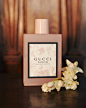 In a close-up, a square-shaped opaque pink glass bottle of Gucci Bloom, eau de toilette, with its black-framed label and pink-on-white delicate floral pattern, stands on a stone surface next to a bunch of ivory colored jasmine blossoms. Out of focus in th