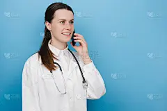 年轻的职业女医生穿着白色的医疗制服，带着听诊器在用手机通话，在蓝色的工作室背景墙上摆姿势。医生谈话。