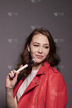 迷人的黑发女子在红色皮夹克与化妆刷孤立在灰色的墙壁背景。专业化妆师
