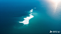 一个从高处俯瞰的海洋场景，广阔的海洋呈现出蓝绿色，波光粼粼。在海洋的中央偏左位置，有一个由海浪形成的白色图形图案，图形边缘清晰可见，仿佛被海浪轻轻勾勒出来。天空是湛蓝的，几朵白云点缀其中，阳光从右侧照