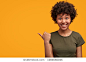 Positive attractive young female with Afro hairstyle, points aside with cheerful expression, shows something amazing at blank space, isolated over bright yellow backgroud. Advertisement and ethnicity