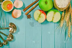 面包店的背景包括苹果，面粉，鸡蛋，肉桂笔记本和铅笔在蓝色的木头桌子上有拷贝空间。在木桌上准备烤苹果蛋