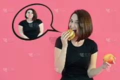 一个苗条漂亮的女孩穿着黑色t恤吃汉堡包，认为她会变胖。一个微笑的女孩拿着一个苹果和一个汉堡在粉红色的