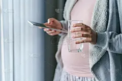 随手拿着陌生白种妇女怀孕母亲的水杯和数码平板，而站在窗口-怀孕和饮水的概念