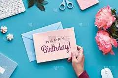 生日贺卡设计。手拿笔，家庭办公。文字生日快乐纸卡片。粉红色牡丹花在蓝色薄荷色表。家庭办公，笔记本，键