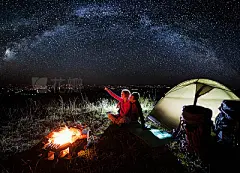 Night camping near the town. Young couple enjoying near campfire and tent, looking at beautiful night sky full of stars and enjoying night scene. Woman is pointing at the sky. Astrophotography