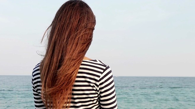长发的年轻女子穿着条纹衬衫看着大海，从后...