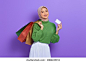 Cheerful beautiful Asian woman in green sweater holding shopping bags and credit card isolated over purple background 库存照片