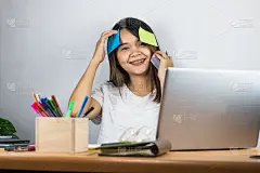 亚洲女性穿白色t恤。在家里快乐的工作眼睛看着相机和有一个灰色的笔记本电脑放在桌子上，手拿着信纸粘在头
