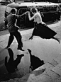 A man gives a woman a helping hand as she takes a flying leap over a large puddle on the pavement. (Photo by Keystone/Getty Images). 1960