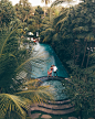 A drone shot of a couple standing between a beautiful round pool and a long pool. She wears a red dess.