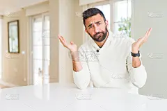 英俊的西班牙裔男子穿着休闲白毛衣在家笨笨和困惑的表情与胳膊和手举起。怀疑的概念。