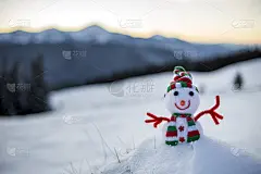 小可爱的玩具娃娃雪人在编织的帽子和围巾在深雪户外在模糊的雪山景观背景。新年快乐，圣诞快乐的贺卡主题。