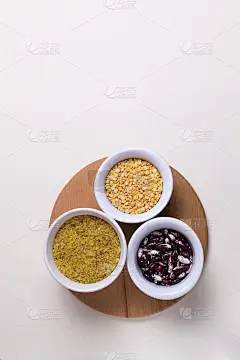 木质砧板上的白色碗里装着碎碎的干小麦、豆子、豌豆，背景为淡黄色。前视图。副本的空间。