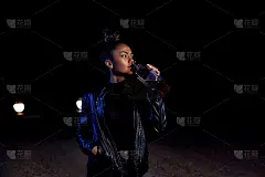 一个穿着皮夹克和黑色衣服的年轻女孩正走在一条废弃的沙路上，手里拿着一瓶威士忌。晚上在灯光下的汽车前灯